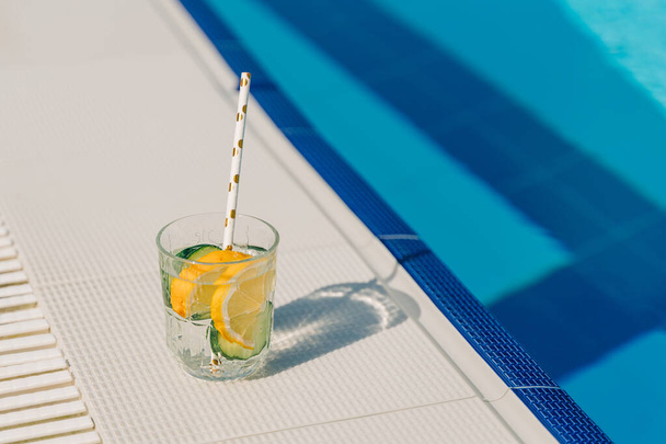 Tropical sparkling cocktail by the pool. The picture of glass with lemon and cucumber lemonade fruit cocktail standing near the poolside. Summer alcohol free drink. Hello summer holiday vacation - Foto, immagini