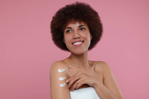 Beautiful young woman applying body cream onto arm on pink background - 写真・画像