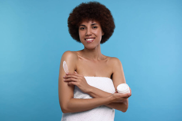 Beautiful young woman applying body cream onto arm on light blue background - Фото, зображення