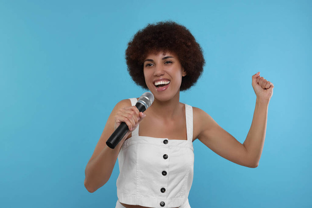 Curly young woman with microphone singing on light blue background - Valokuva, kuva