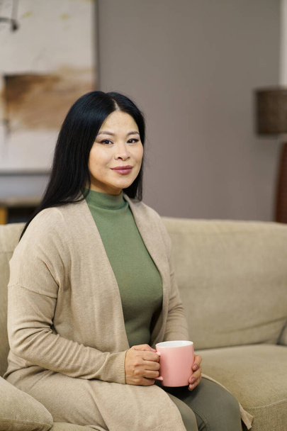 Cute Asian MILF sitting on couch with cup of coffee home. The image captures moment of relaxation and leisure as woman enjoys her coffee break in comfort of her own home. . High quality photo - Fotografie, Obrázek