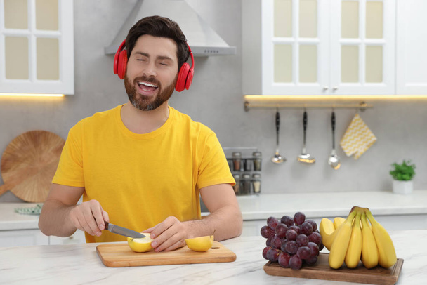 Handsome man listening music with headphones while cutting apple at white marble table in kitchen. Space for text - Valokuva, kuva