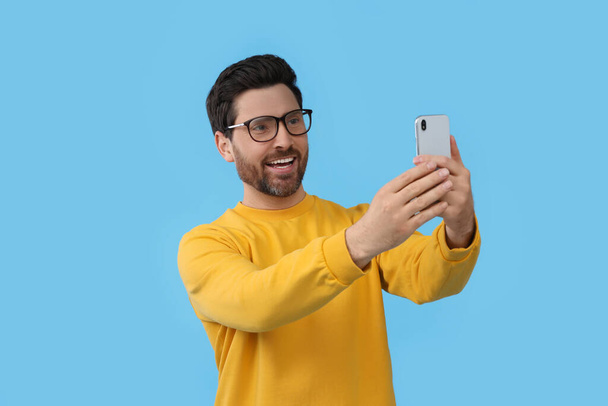 Smiling man taking selfie with smartphone on light blue background - Zdjęcie, obraz