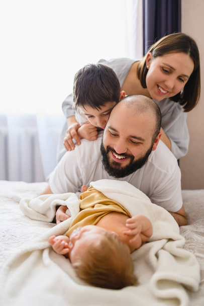 Happy family of four with newborn baby in home bed, mother, father and older brother play with infant - Zdjęcie, obraz