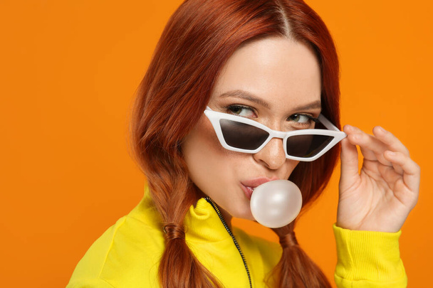 Portrait of beautiful woman in sunglasses blowing bubble gum on orange background - Photo, Image