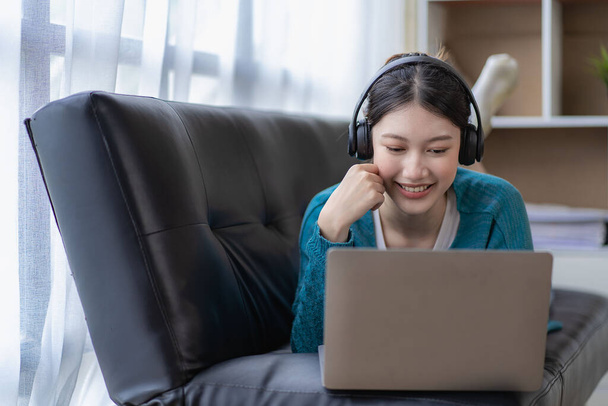 Jonge mooie Aziatische vrouw in casual kleding zitten op de bank met behulp van computer laptop vermakelijke sociale media om te ontspannen, glimlachen en lachen. - Foto, afbeelding