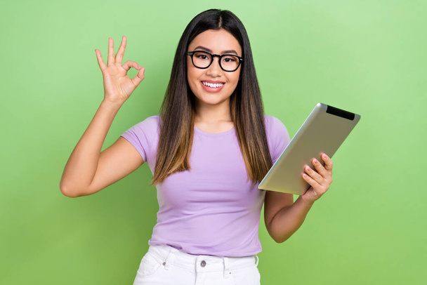 Photo of lovely young vietnamese chinese lady hold tablet show okey specs dressed stylish violet garment isolated on green color background. - Photo, image