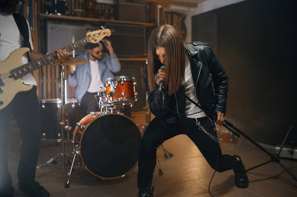 Εκφραστικός ροκάς τραγουδά σε μικρόφωνο ενώ η μπάντα του παίζει heavy metal σε drum και ηλεκτρική μπάσα κιθάρα. Νέοι στην επανάληψη πρόβας στο στούντιο - Φωτογραφία, εικόνα