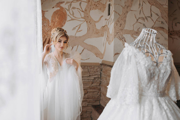 Una sposa in vestaglia posa dietro un manichino con un abito da sposa sullo sfondo di pareti dipinte. - Foto, immagini