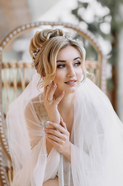 Eine blonde Braut sitzt in einem Holzstuhl, sie trägt ein Gewand und einen langen Schleier. Hochzeitsportrait Hochzeitsfoto in einem leichten Schlüssel. Schönes Make-up und Haare. Der Morgen der Braut - Foto, Bild