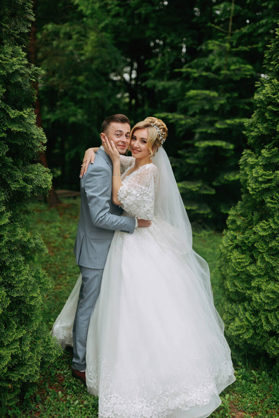 Portrait of the bride and groom standing on the background of green trees, embracing. The bride smiles sincerely. Stylish groom. Fashion and style. Beautiful bride - Photo, image