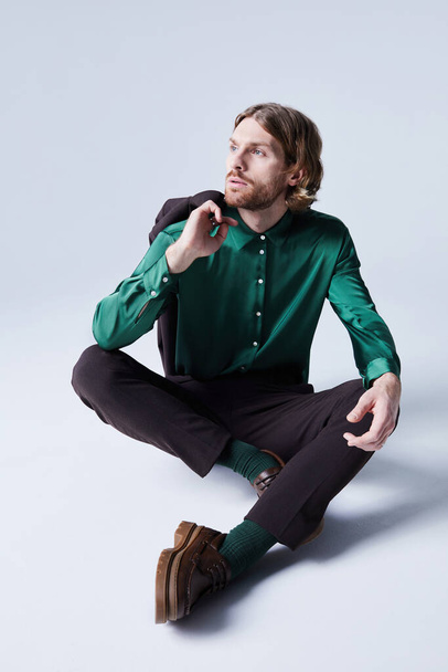 Trend portrait of male fashion model posing casually wearing suit with teal green silk shirt and sitting on floor - Фото, зображення