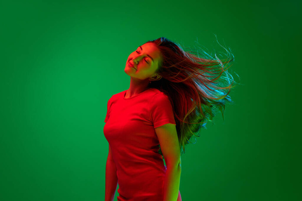 Portré gyönyörű, fiatal, barna lány piros ruhában pózol zöld háttér neon fény. Gyengédség és nőiesség. Fogalom a fiatalok, emberi érzelmek, életmód, arckifejezés, reklám - Fotó, kép