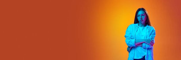 Ritratto di giovane donna in abiti casual in piedi con volto offeso sullo sfondo dello studio arancione sfumato in luce al neon. Concetto di gioventù, emozioni umane, stile di vita. Bandiera. Copia spazio per l'annuncio - Foto, immagini