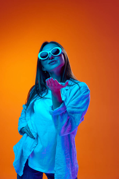 Portrait of young woman in casual clothes posing in trendy sunglasses against gradient orange studio background in neon light. Concept of youth, human emotions, lifestyle, facial expression, ad - Φωτογραφία, εικόνα