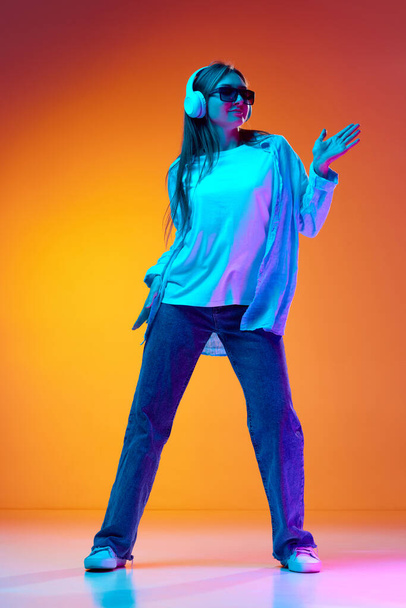 Jovem mulher em roupas casuais e óculos de sol ouvindo música em fones de ouvido e dançando contra gradiente de fundo estúdio laranja em luz de néon. Conceito de juventude, emoções, estilo de vida, festa, anúncio - Foto, Imagem