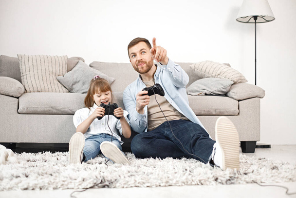 Loving father playing with his daughter with Down syndrome at home together. Man and girl sitting on a floor near sofa and holding joysticks. Bearded man wearing blue shirt and girl white shirt. - Fotó, kép