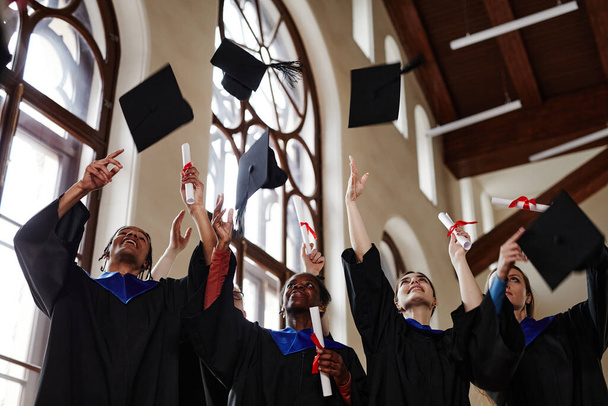 Groupe de jeunes gens joyeux portant des robes de remise des diplômes lors d'une cérémonie à l'université et jetant des casquettes dans l'air - Photo, image