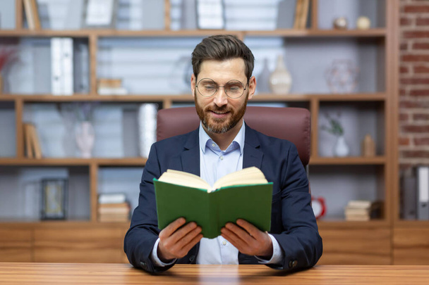Portrait of a young businessman reading a book in the office. The teacher teaches students dissonantly through a video call, gives a lecture. A pastor, a priest reads a sermon from the Bible online. - Фото, изображение