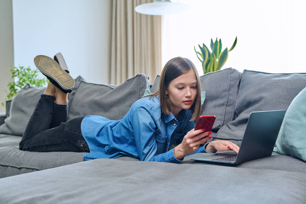 Young smiling happy woman using smartphone laptop, lying on sofa at home in living room. Internet online services, communication, mobile applications, technologies for leisure study work lifestyle - Фото, зображення