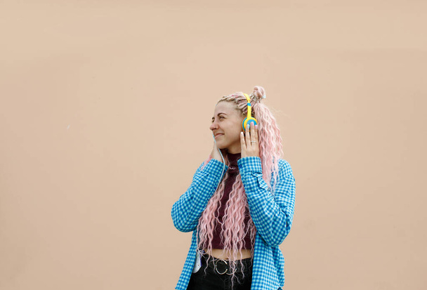 Una giovane donna felice con i capelli lunghi rosa e una camicia blu. Una ragazza con le cuffie su uno sfondo luminoso. Spazio vuoto per testo pubblicitario. - Foto, immagini