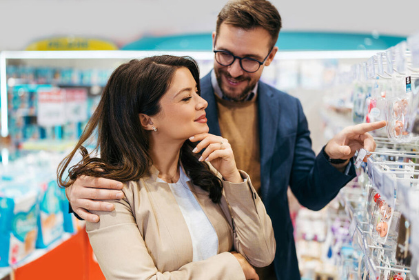 Attractive middle age couple enjoying in buying clothes and appliances for their new baby. Heterosexual couple in baby shop or store. Expecting baby concept. - Photo, image