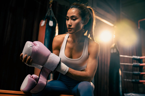 Határozott ázsiai nő Muay Thai bokszoló izomzattal fizikai felkészültség test burkolja a kezét és a dons vagy visel boksz kesztyű, felkészülés intenzív boksz edzés a ringben az edzőteremben. Impetusz - Fotó, kép