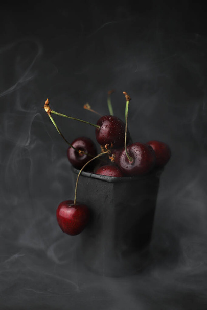 летние сладкие спелые вишни красно-бордового цвета в черном стакане на черном фоне в белом дыму. для открыток, листовок, баннеров, этикеток, заставки и многое другое - Фото, изображение