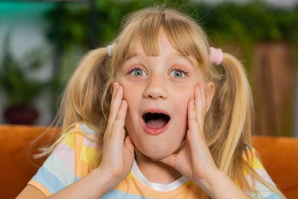 Oh my God, Wow. Caucasian surprised preteen school girl looking at camera with big eyes, shocked by sudden victory, good win news, celebrating. Excited amazed child kid at home room sitting on couch - Fotografie, Obrázek