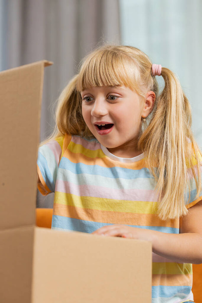 Happy child kid girl unpacking delivery parcel. Smiling satisfied teen toddler shopper online shop customer opening cardboard box receiving purchase gift by fast postal shipping at home. Vertical shot - Photo, image