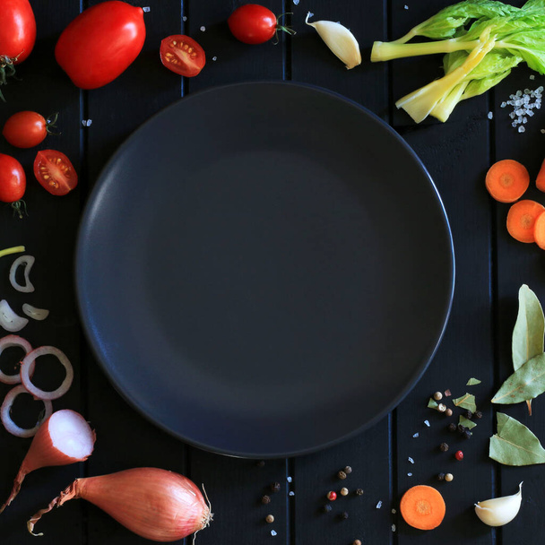 Gesunde Ernährung oder Ernährungskonzept. Gemüse, Gewürze und Kräuter um einen leeren Teller herum auf einem schwarzen Holztisch. Kopierraum. Gesamtübersicht. - Foto, Bild
