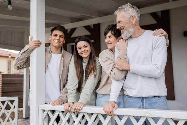 parents day, middle aged parents hugging and looking away near teenage daughter and adult son on porch of summer house, family celebration, bonding, modern parenting, translation of tattoo: harm none - Photo, Image