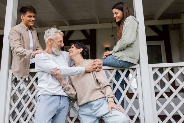 Ebeveynler günü, neşeli kardeşler orta yaşlı ebeveynlere bakıyor. Yazlık evin verandasında sarılıp gülümsüyorlar. Aile kutlaması, bağlanma, hatırlanacak anlar, modern ebeveynlik, mutlu evlilik. - Fotoğraf, Görsel