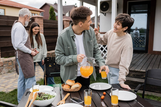 celebration of parents day, modern parenting, happy middle aged mother talking to young adult son with jug of orange juice, father and daughter preparing food on bbq grill, summer, backyard  - Photo, Image