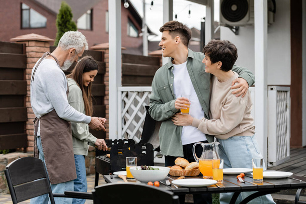 modern parenting, happy parents day, middle aged mother hugging with cheerful young adult son holding glass of orange juice, father and teenage daughter preparing food on bbq grill, summer, backyard  - Photo, Image