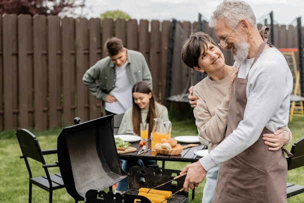 parents day, middle aged couple smiling during family bbq party, bearded tattooed man holding tongs near bbq grill, grilling corn, daughter and son, translation of tattoo: harm none do what you will  - Photo, Image
