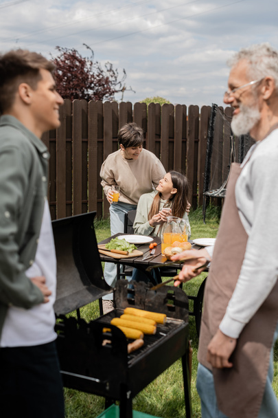 cheerful teenage girl holding smartphone and looking at mother digital age, father and son preparing food on bbq grill, barbecue party, parents day cerebration, backyard, blurred, candid  - Fotografie, Obrázek