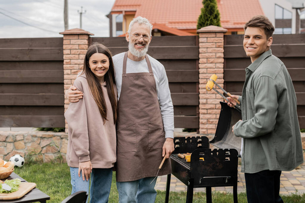 happy parents day, middle aged father hugging teenage daughter, young adult son holding tongs and grilling corn, bbq party, family photo, looking at camera, backyard of summer house  - Foto, imagen