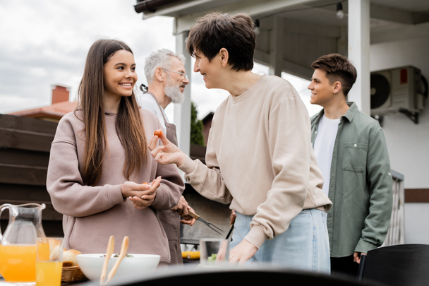 happy middle aged mother holding cherry tomato and talking with teenage daughter, family grill party, father and son on blurred background, happy parents day concept, summer - Photo, Image