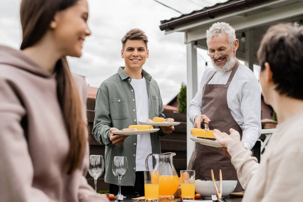 bearded father and his young adult son holding plates with grilled corn during bbq party, young adult son looking at mother and teenage sister on blurred foreground, backyard  - Foto, Bild