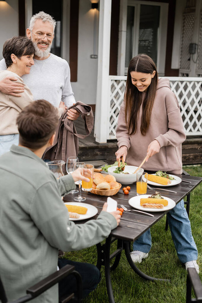 celebrating parents day, cheerful middle aged couple hugging near joyful teenage girl serving salad next to adult brother with glass of orange juice, love, family grill party, summer, happy marriage  - 写真・画像