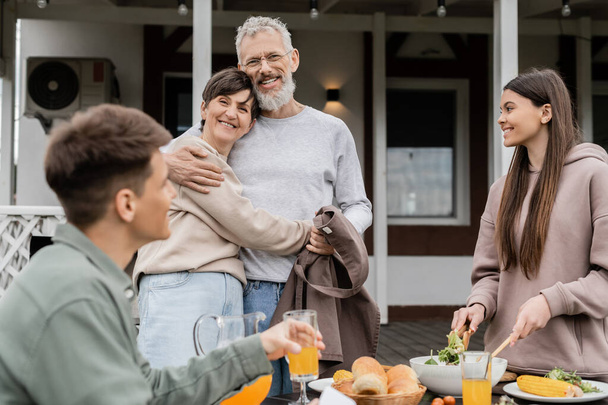 happy marriage, celebrating parents day, cheerful middle aged couple hugging near joyful teenage daughter mixing salad next to adult brother, love, family grill party, summer,  summer house backyard  - Photo, Image