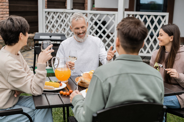 весёлый мужчина средних лет смотрит на взрослого сына во время семейной барбекю-вечеринки, сидит на заднем дворе летнего домика, проводит время вместе, ест гриль барбекю, счастливые родители день концепции  - Фото, изображение