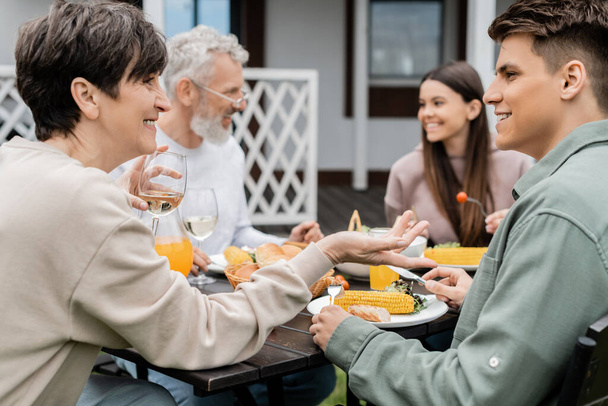 happy middle aged mother holding glass of wine and talking with adult son during bbq family party, eating grilled food, father and daughter on blurred background, backyard of summer house  - Photo, Image