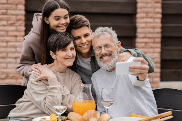 Smiling middle aged father taking selfie on smartphone with family and kids near tasty summer food during bbq party and parents day celebration at backyard, family love and unity concept - Фото, зображення