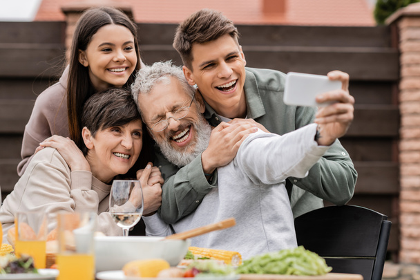 Iloiset lapset halaavat keski-ikäisiä vanhempia samalla, kun he ottavat selfien älypuhelimella yhdessä grillijuhlien aikana ja vanhemmat juhlivat takapihalla kesäkuussa, onnelliset vanhemmat päivä konsepti - Valokuva, kuva