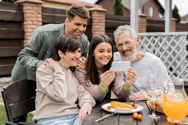 Smiling teen girl using smartphone near middle aged parents and brother during barbeque party with summer food and parents day celebration at backyard in june, happy parents day concept - Photo, Image