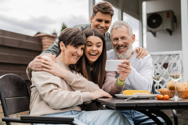 Cheerful teenage girl holding smartphone near middle aged parents and young brother during bbq summer party and parents day celebration at backyard in june, happy parents day concept - Photo, Image