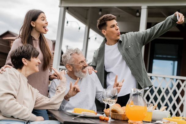 Young man sticking out tongue while taking selfie with sister and cheerful middle aged parents during bbq party and parents day celebration at backyard in june, happy parents day concept - Photo, Image