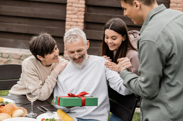 Mosolygó középkorú férfi kezében ajándék doboz közelében a gyermekek és a feleség alatt grillparti nyári étel és a szülők napi ünneplés a kertben júniusban, ünnepli szülői nap koncepció - Fotó, kép